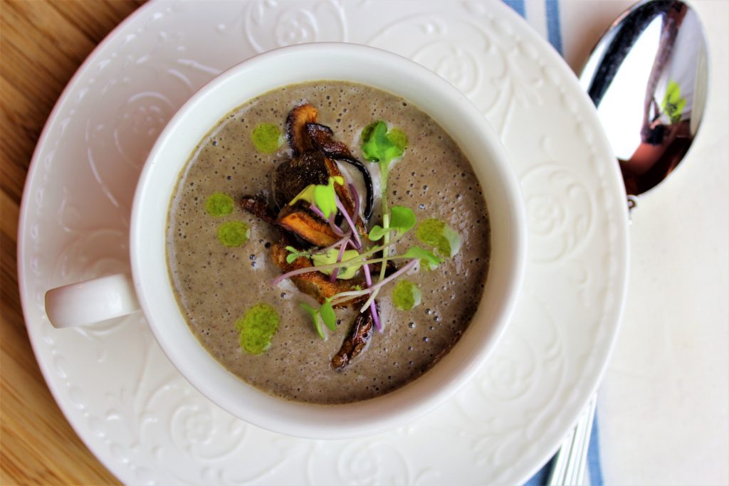 an elegant white coffee cup and saucer filled with mushroom soup, crispy mushrooms and a spoon and cloth napkin.